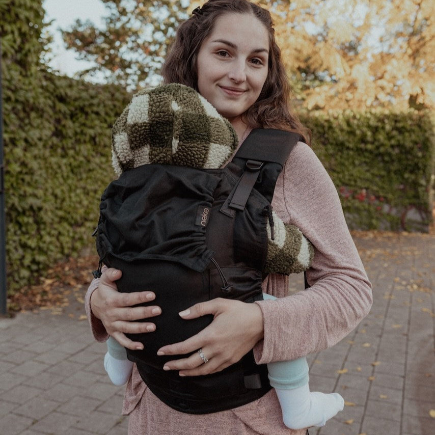 Woman carrying a baby in a carrier outdoors with trees and pavement in the background