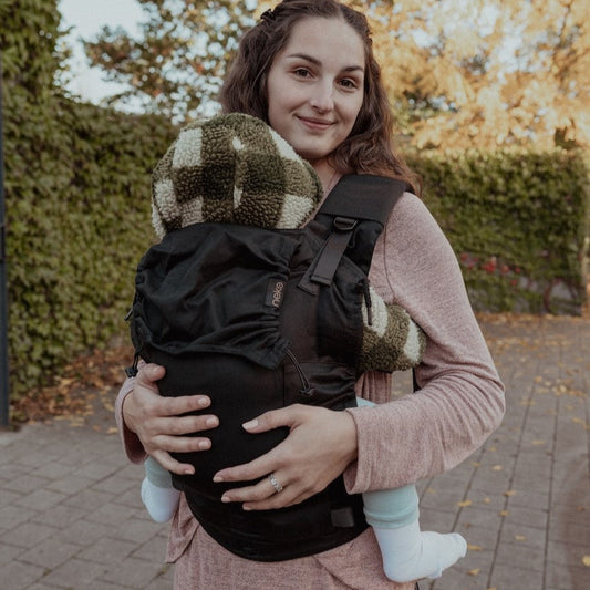 Woman carrying a baby in a carrier outdoors with trees and pavement in the background
