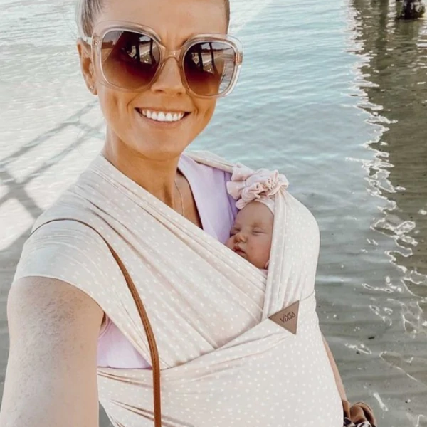 A person wearing sunglasses is smiling while carrying a sleeping infant in a beige stretch wrap baby carrier.