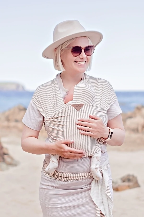Smiling woman wearing a striped stretch wrap baby carrier near the ocean