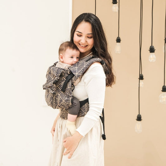 A baby in a geometric patterned baby carrier with dark blue and beige stripes being worn by a person with long brown hair.