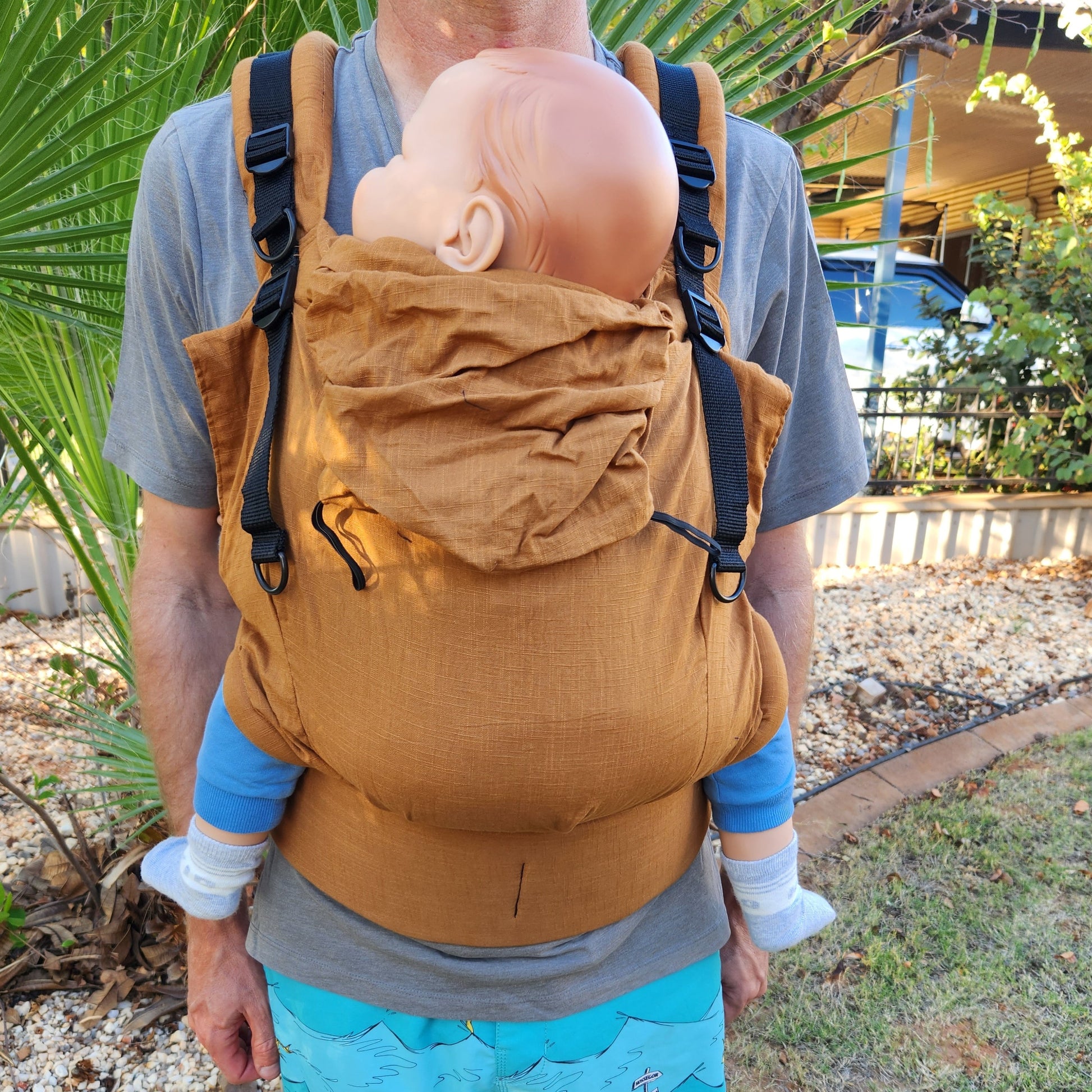 Toddler sized babywearing doll in sandy, rusty coloured full buckle carrier, worn by a man