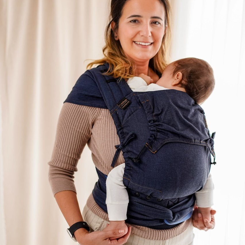 Woman holding a baby in a navy blue baby carrier against a beige curtain background
