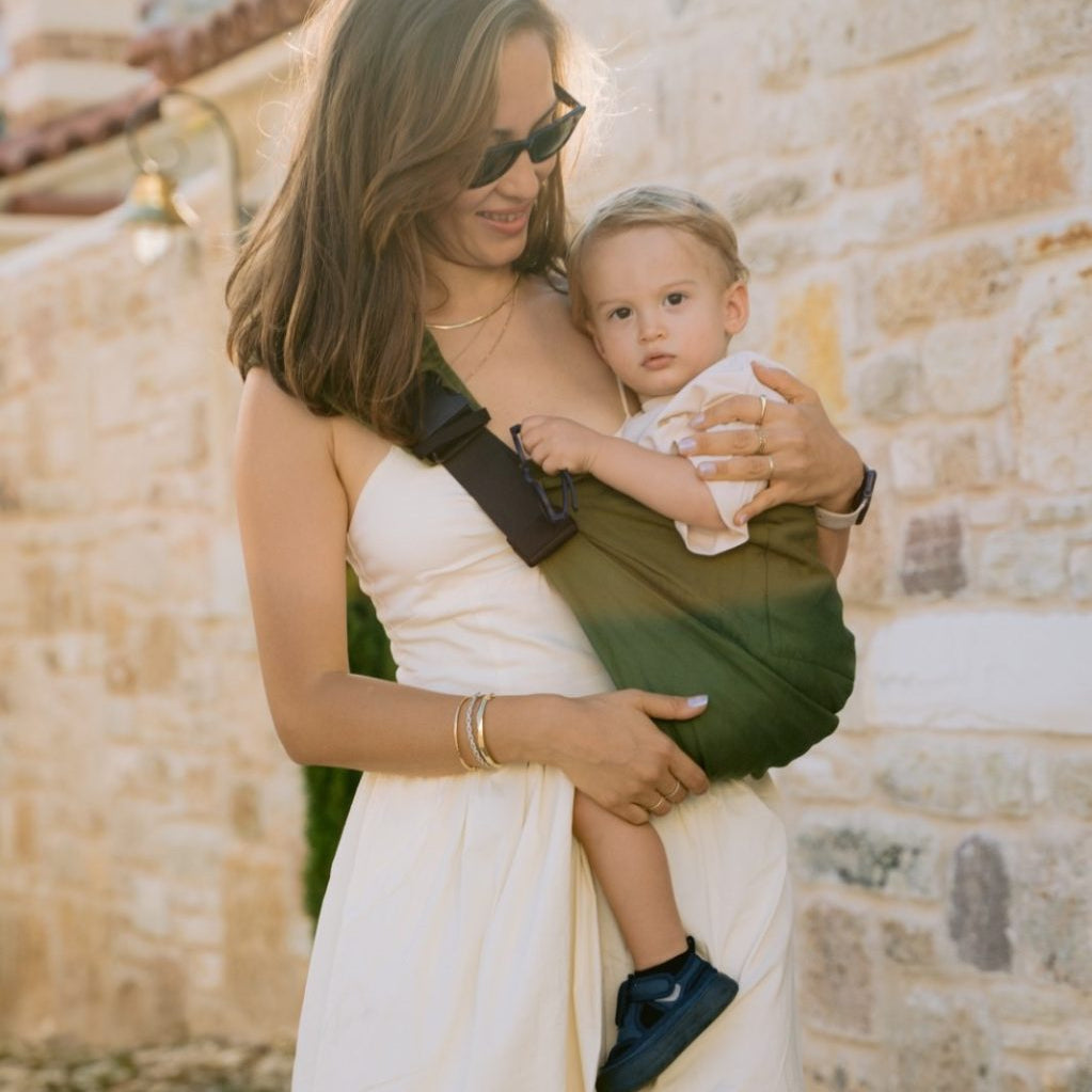 Woman in a white dress holding a baby in a green sling against a stone wall.
