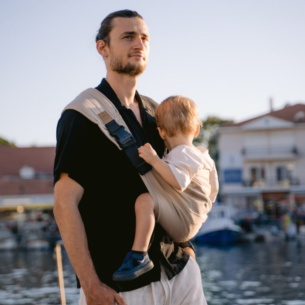 Man holding a child in a sling by a waterfront with boats and buildings in the background