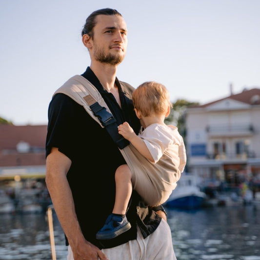 Man holding a child in a sling by a waterfront with boats and buildings in the background