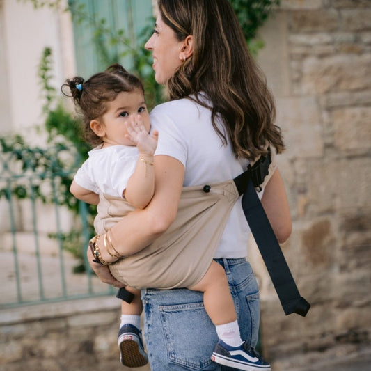 Woman carrying a child in a Neko sling outdoors with stone wall and green door in the background