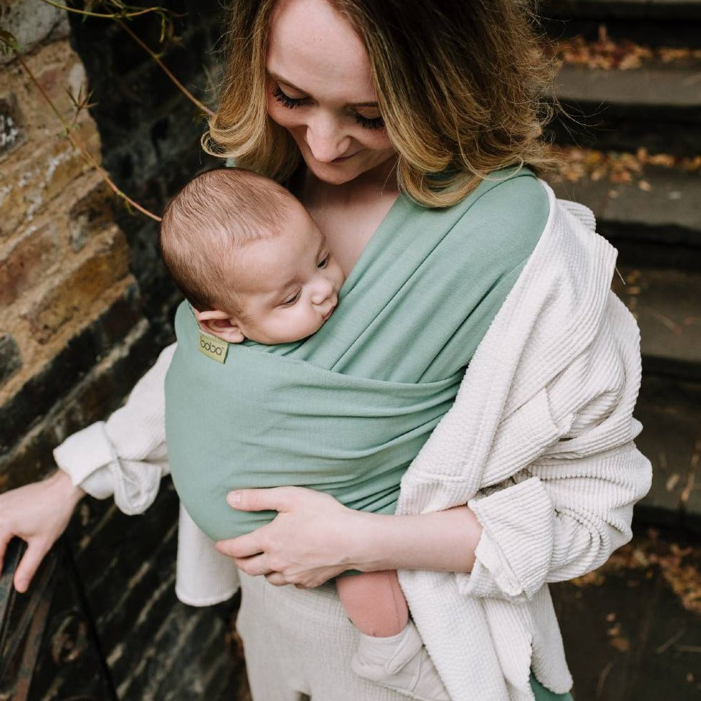 Mother standing on concrete steps has baby in light green stretch wrap. Baby is looking out over their shoulder